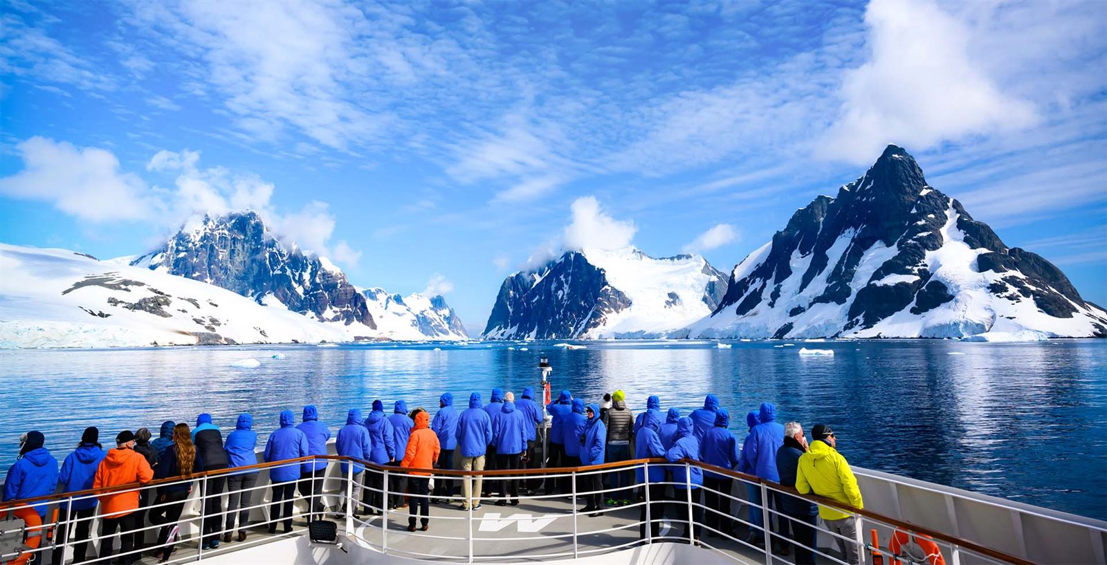 Expedition cruise passengers in blue parkas on deck, facing snow-capped Antarctic peaks