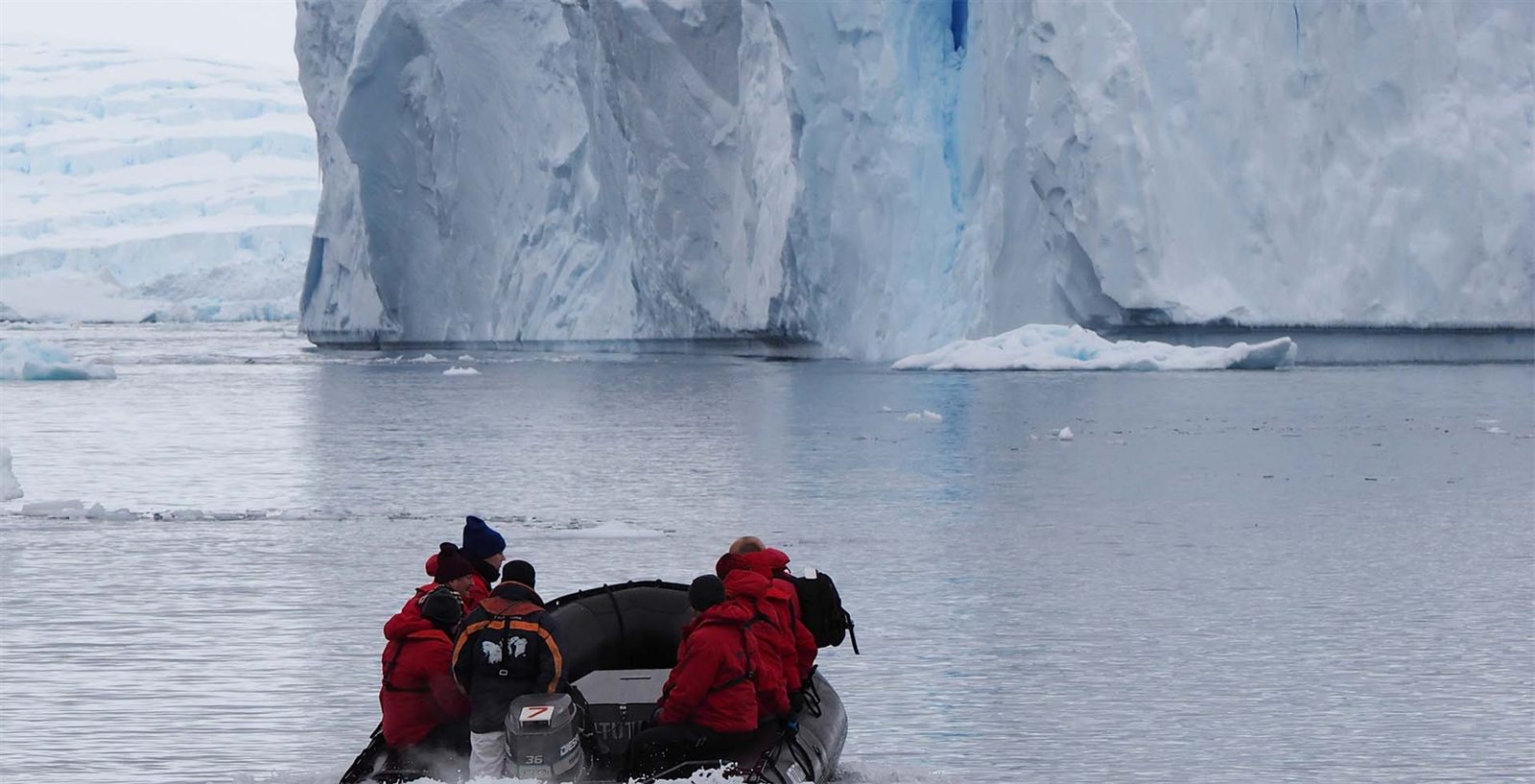 Zodiac with expedition guests in red parkas below a towering Antarctic ice cliff